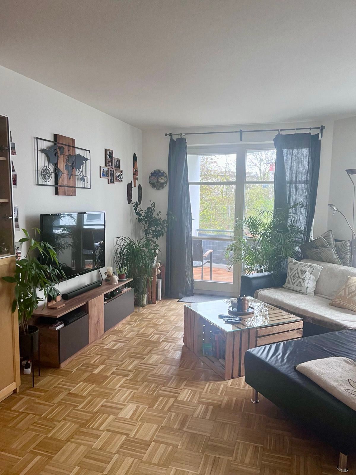 A cozy living area is depicted with a combination of wooden furniture and plants. A large window allows natural light to illuminate the space, revealing a comfortable sofa and a decorative coffee table. Artwork adorns the walls, enhancing the inviting atmosphere.