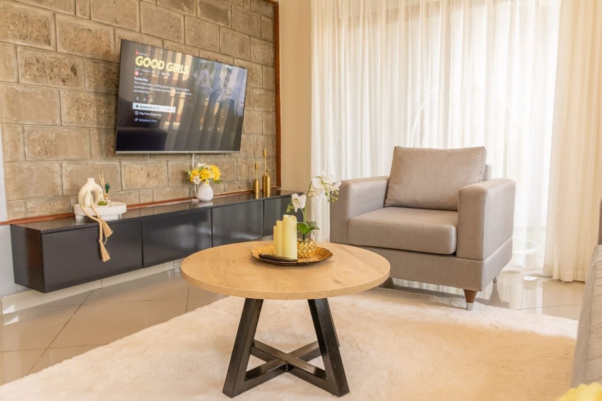 A modern living area is featured with a large flat-screen TV mounted on a textured stone wall. A light-toned wooden coffee table sits on a soft area rug, complemented by a gray armchair. Natural light filters through sheer curtains, enhancing the space's brightness.
