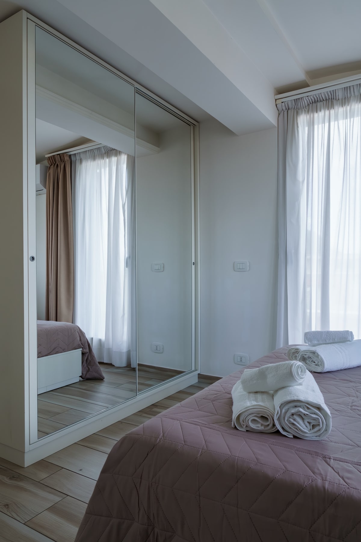 The bedroom features a spacious bed covered in a subtle quilt. Neatly rolled towels are arranged at the foot of the bed. A large mirrored wardrobe reflects natural light flowing in through the white curtains. Soft, neutral tones create a calm environment.