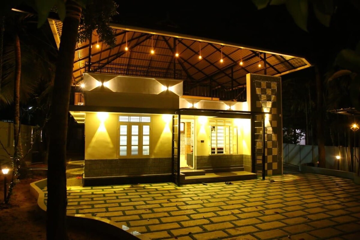 The exterior of a modern house is shown at night, featuring a well-lit entrance with glass panel doors. A spacious patio area is highlighted by warm yellow lights, while decorative wall accents and a stone pathway create an inviting atmosphere.
