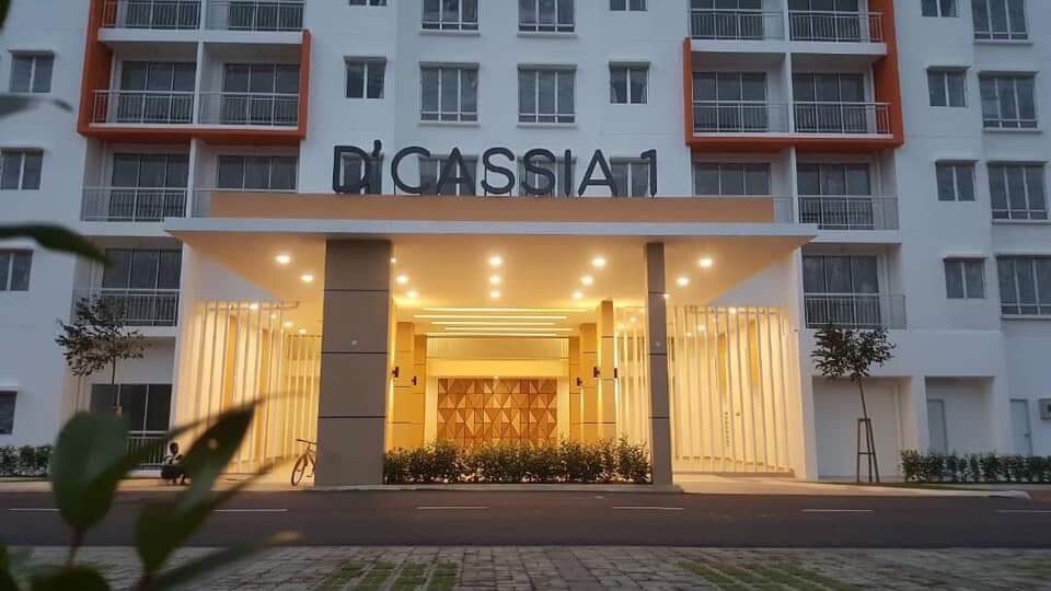 A modern building entrance is highlighted by warm, ambient lighting. The tall structure features a distinctive geometric wooden panel, alongside symmetrical plants that frame the pathway. Multiple balconies are visible on the upper floors, enhancing the contemporary aesthetic.