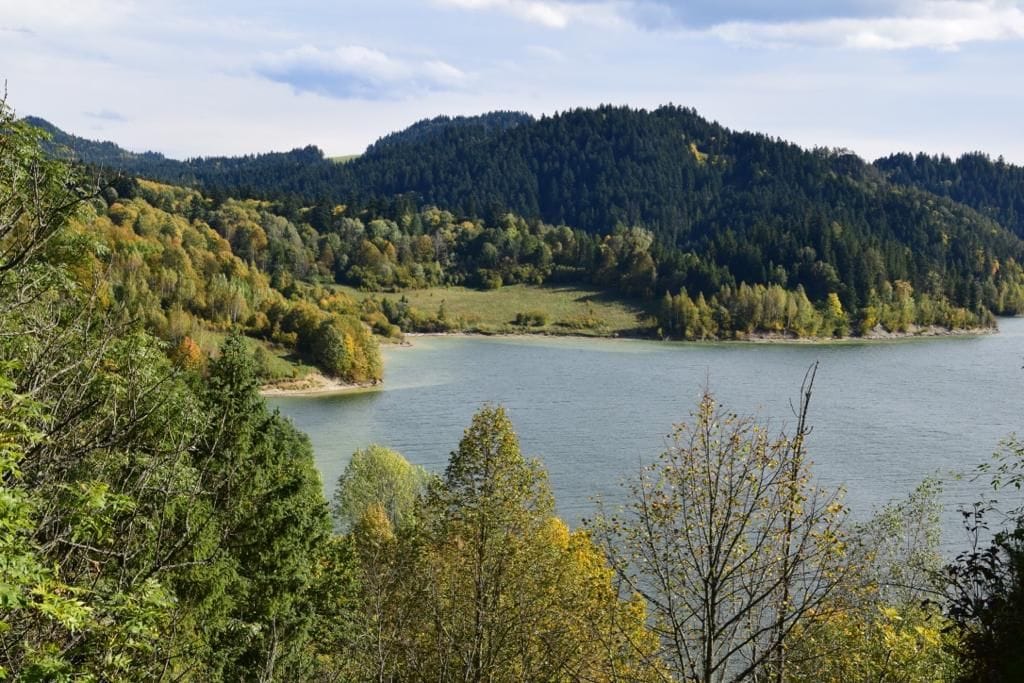A serene landscape view showcases a calm lake surrounded by dense greenery and rolling hills. Trees display a range of autumn colors, with golds and greens blending harmoniously. The peaceful water reflects the natural surroundings under a partly cloudy sky.