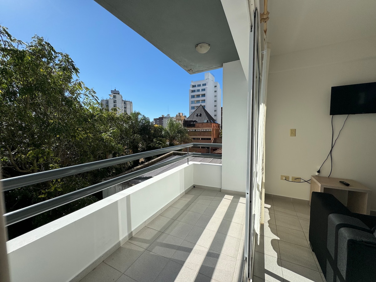 A sunny balcony offers views of the tree-lined street and nearby buildings. The space features a tiled floor, with a railing running along its perimeter. A wall-mounted television is visible inside the room, enhancing the open and airy feel.
