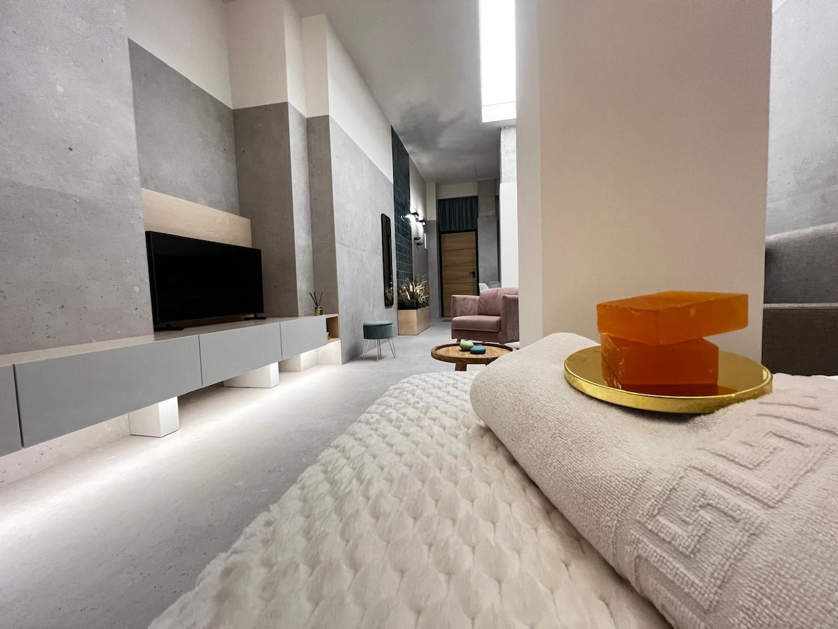 A modern living area is seen from a cozy vantage point, showcasing a low television unit against textured walls. A soft blanket and decorative items are placed on a tray beside a stack of orange soap bars, with soft lighting enhancing the serene ambiance.