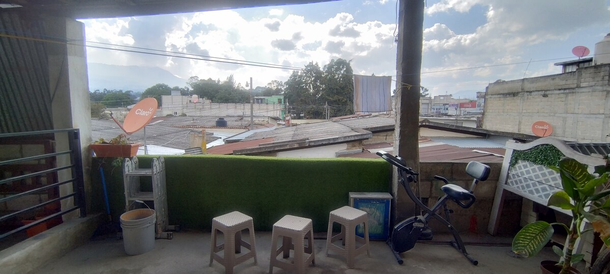 A balcony area features three lightweight stools arranged on a tiled floor, surrounded by a small green space and potted plants. In the background, rooftops and distant buildings are visible under a partly cloudy sky.