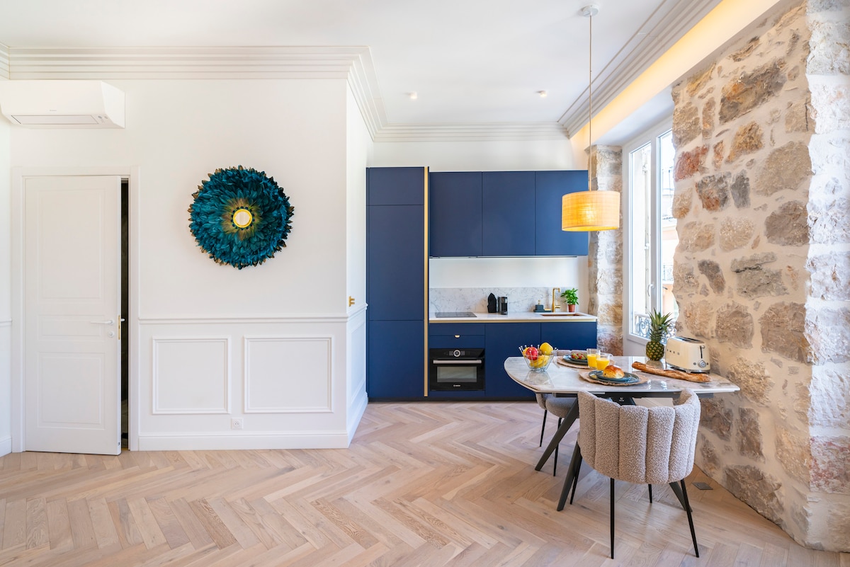 The kitchen and dining area features a circular table set for two, adorned with fresh fruit and a drink. Stone walls add texture, while modern cabinetry in deep blue complements the wood-finish floor. A warm light fixture hangs above, enhancing the inviting space.