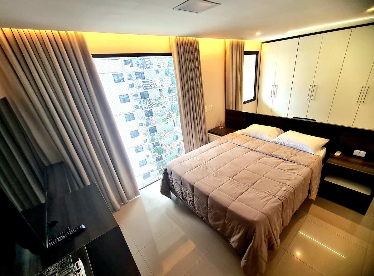 A spacious bedroom features a large bed dressed in a neutral-toned coverlet, complemented by soft lighting. Sheer curtains allow natural light to filter through expansive windows, showcasing views of the city. A wooden desk is positioned nearby, creating a functional workspace.