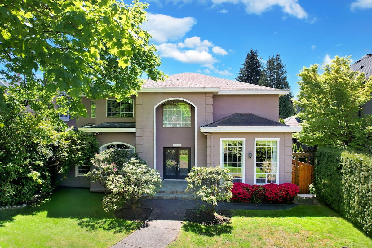 The exterior of the home is showcased with lush greenery and vibrant flowering bushes. A welcoming entrance features a large front door framed by large windows, allowing natural light to illuminate the interior. The two-story structure presents a blend of modern and classic architectural elements.