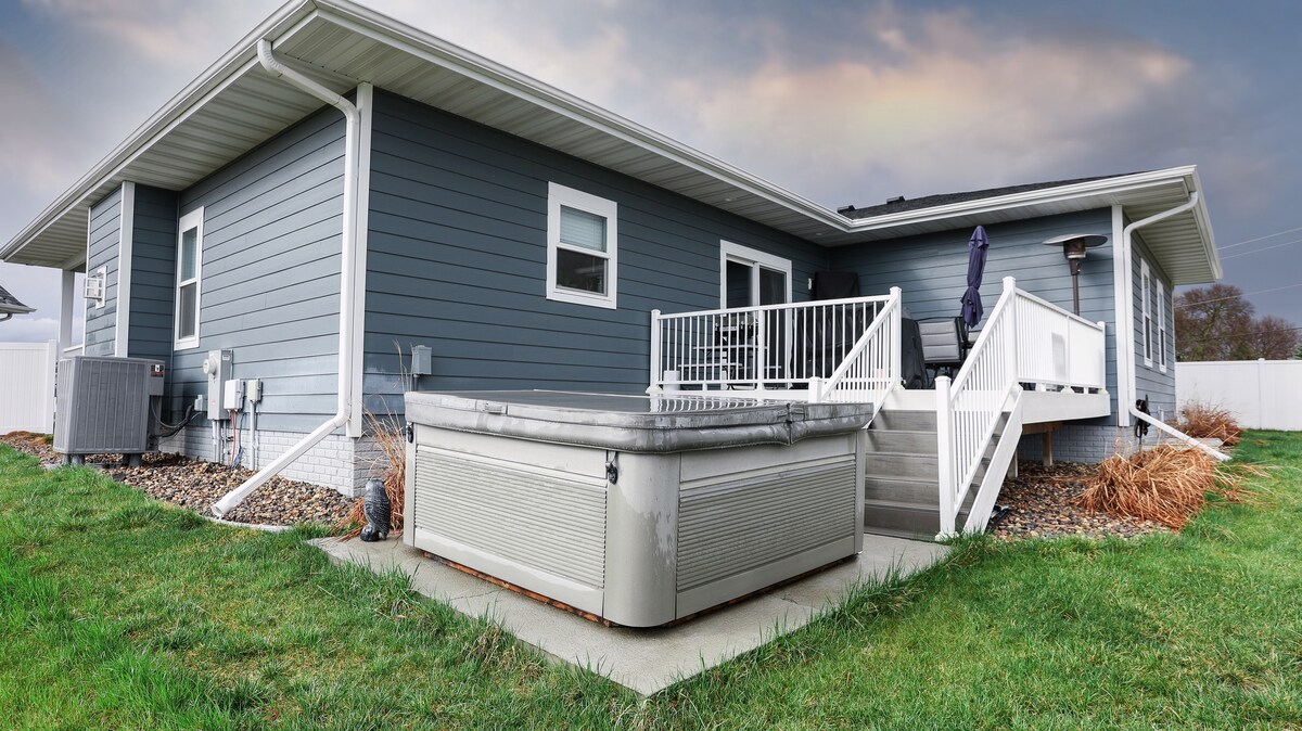 The exterior of a ranch-style home is depicted, featuring gray vinyl siding and a covered deck. A hot tub is positioned on a concrete pad in the foreground, surrounded by grass and landscaping. Steps lead from the deck to the yard, enhancing accessibility.