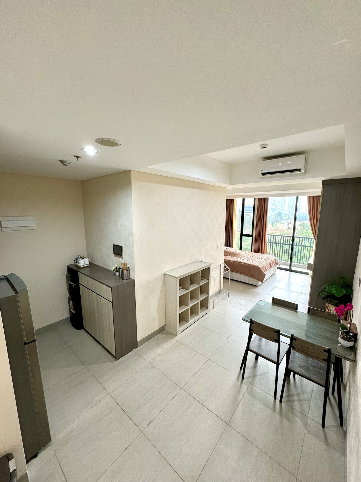 An open-concept living area is shown, featuring a fully equipped kitchen on the left, a dining table with four chairs in the center, and a queen-sized bed in the background near large glass doors leading to a balcony. Natural light fills the space.