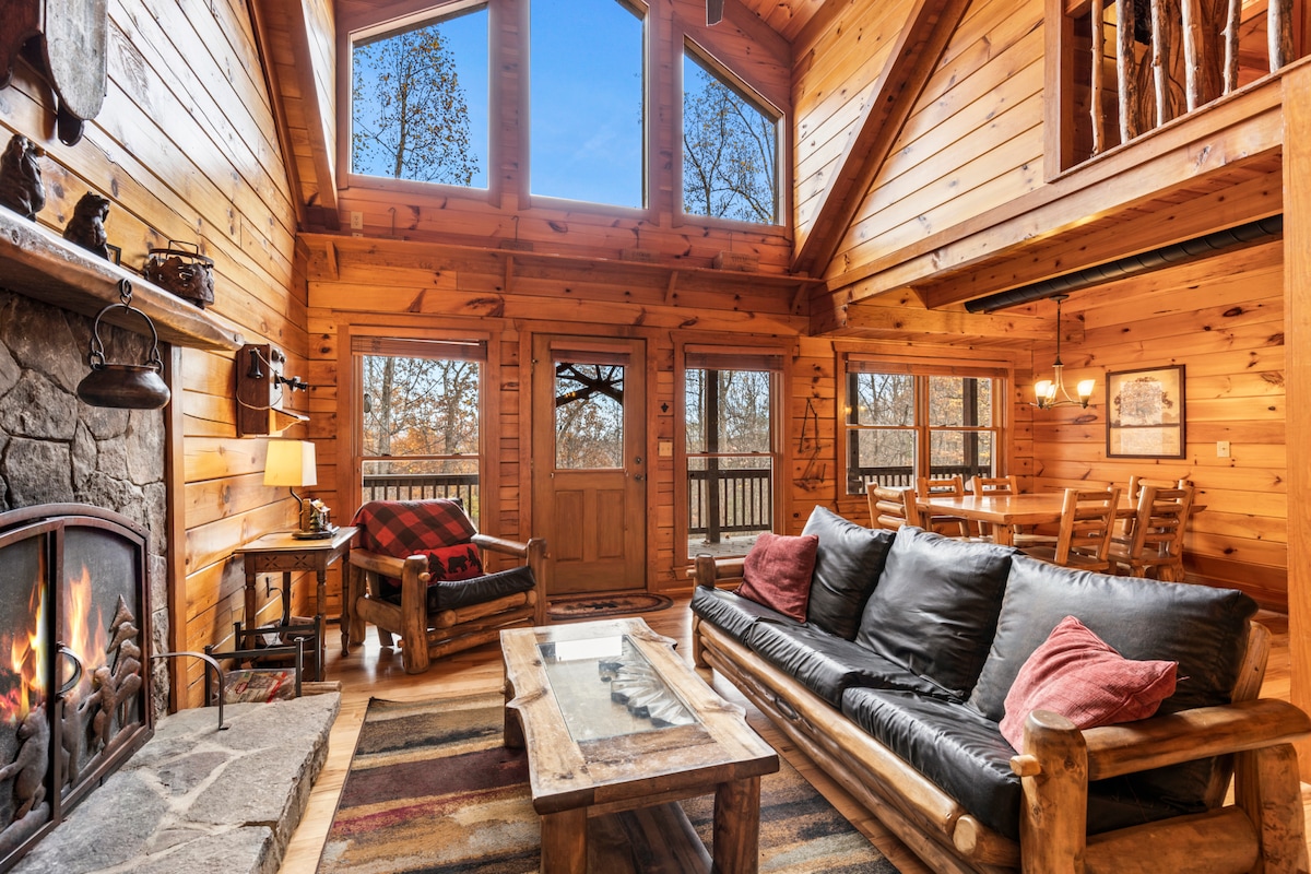 A cozy living area is framed by wooden walls and ceiling. Large windows bring in natural light and showcase outdoor views. A stone fireplace adds warmth, and comfortable seating is arranged around a rustic coffee table. A dining area can be seen in the background.