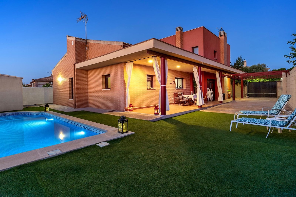 The exterior of a spacious house features a well-lit patio with a seating area, surrounded by lush greenery. A swimming pool glistens nearby, offering a place for relaxation. The structure is enhanced by soft lighting and a tranquil ambiance as evening approaches.