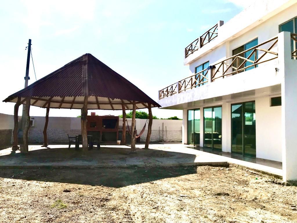 An outdoor area features a shaded gazebo with a thatched roof, accompanied by a table and chairs. The modern structure displays large glass windows and balconies, creating a connection to the surrounding landscape. The ground is spacious and mostly clear, offering ample room for activities.
