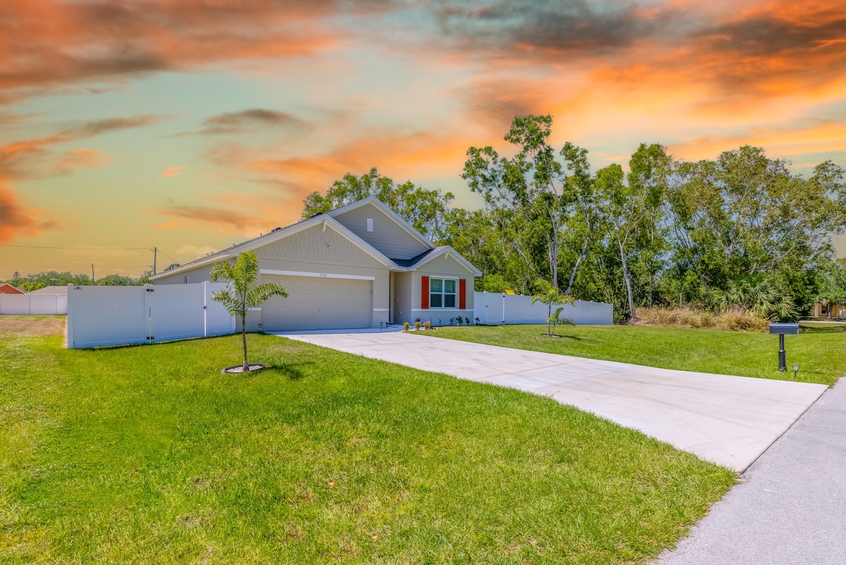 A single-story house with a welcoming facade is set against a colorful sunset sky. The spacious driveway leads to a two-car garage, flanked by green grass and young landscaping, including small trees. A white fence surrounds the property, enhancing its privacy.