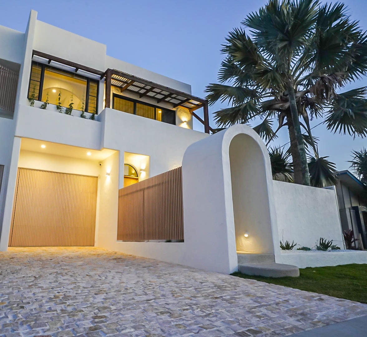 The entrance of the villa showcases a modern architectural design with smooth white walls and warm lighting. A cobblestone pathway leads to the garage, while tropical palm trees and greenery frame the entrance, creating an inviting first impression.
