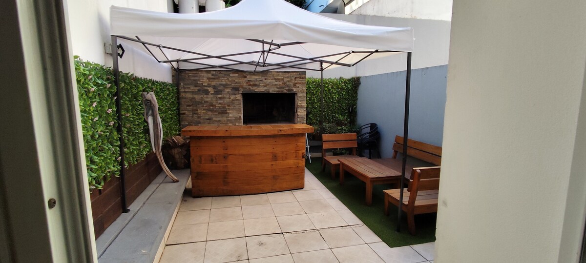 A spacious outdoor patio features a wooden bar area and lounge seating arranged around a stone wall. A white canopy provides shade, while greenery lines the walls, creating a relaxing environment for leisure and social gatherings.