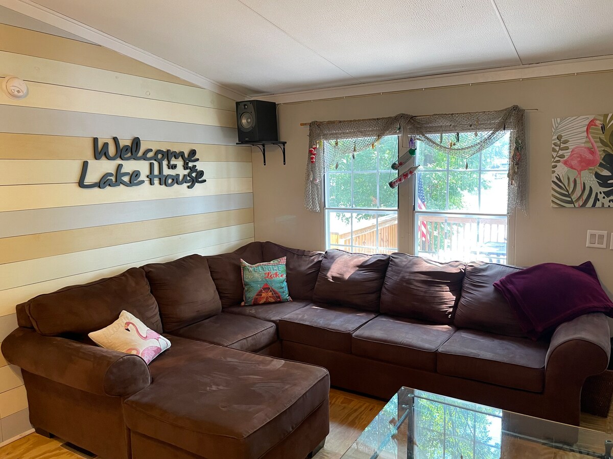 A welcoming living space features a large brown sectional sofa with a chaise, accented by colorful throw pillows. Natural light floods in through the window adorned with light sheer curtains, creating a relaxed ambiance. A decorative sign reading 'Welcome Lake House' adds charm to the room.
