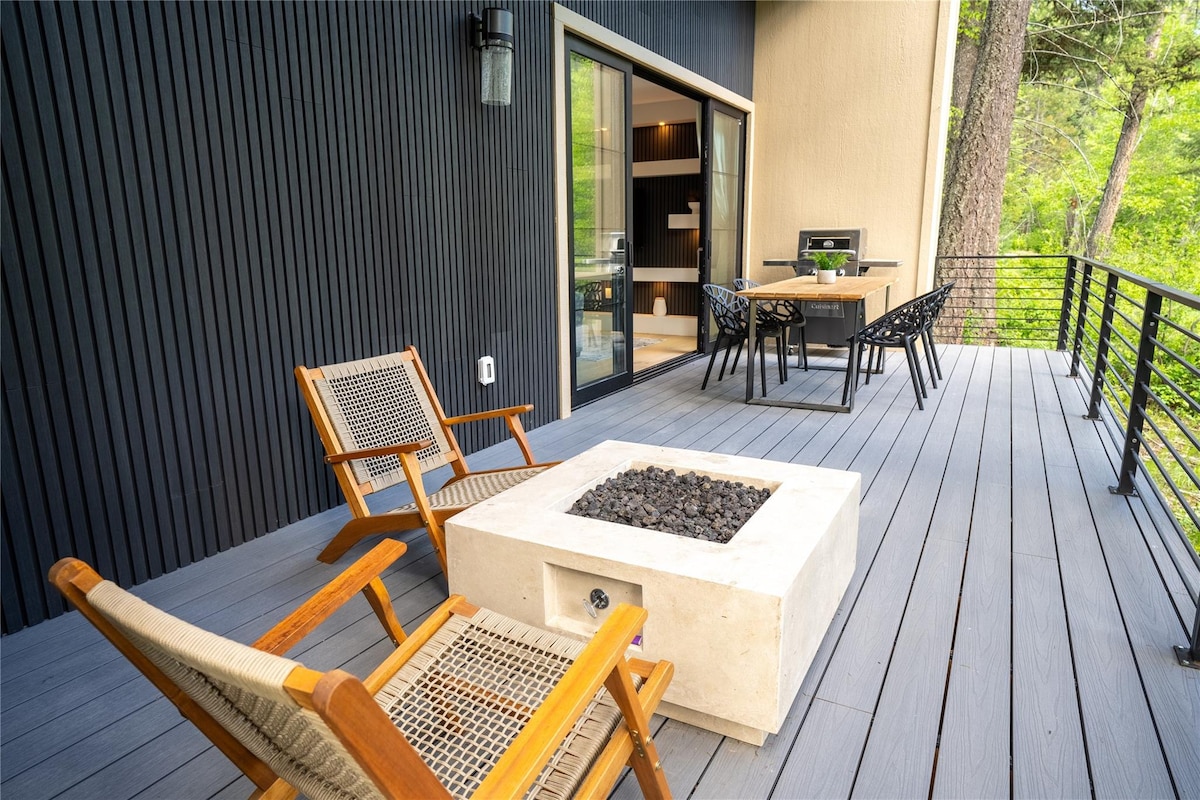 An outdoor patio features a modern fire pit surrounded by two wooden chairs and a dining table with four black chairs. The space is framed by lush greenery, and glass sliding doors lead to the interior, offering a seamless transition between outdoor and indoor living.