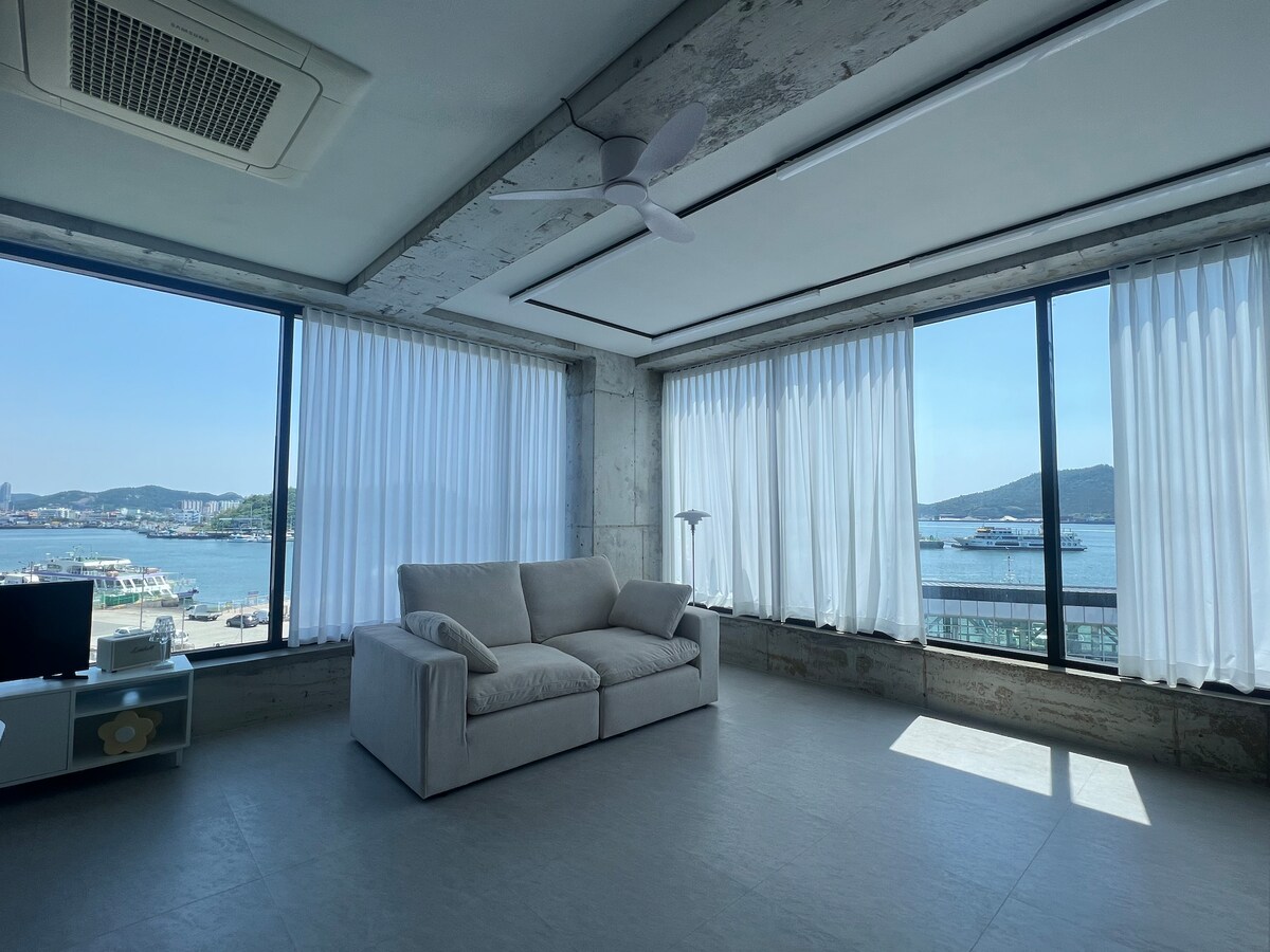 A spacious living area features large windows with sheer curtains, allowing for natural light and views of the harbor. A light-colored sofa is positioned to face the water, creating a comfortable space for relaxation. A ceiling fan is visible above, enhancing airflow.