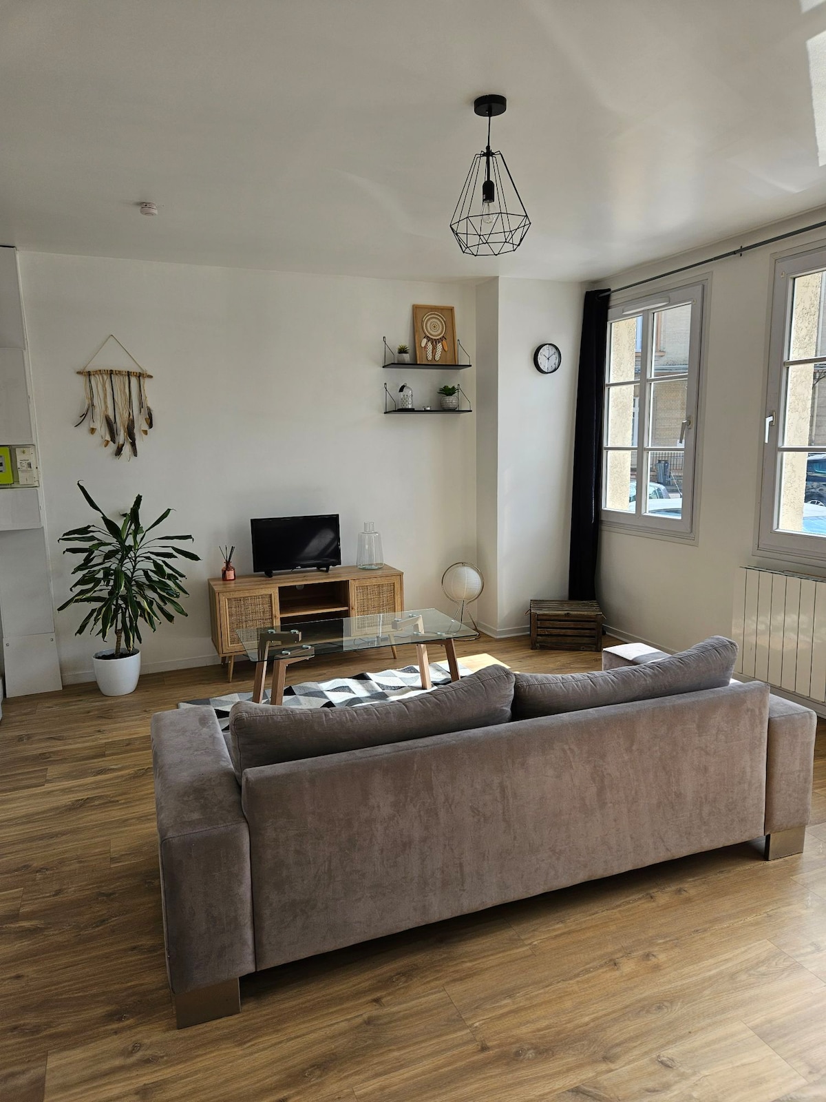 A well-lit living area features a comfortable sofa positioned near a compact entertainment unit. A plant adds a touch of greenery, while a wall-mounted shelf displays decorative items. Natural light enters through large windows, complemented by minimalist decor and a neutral color palette.