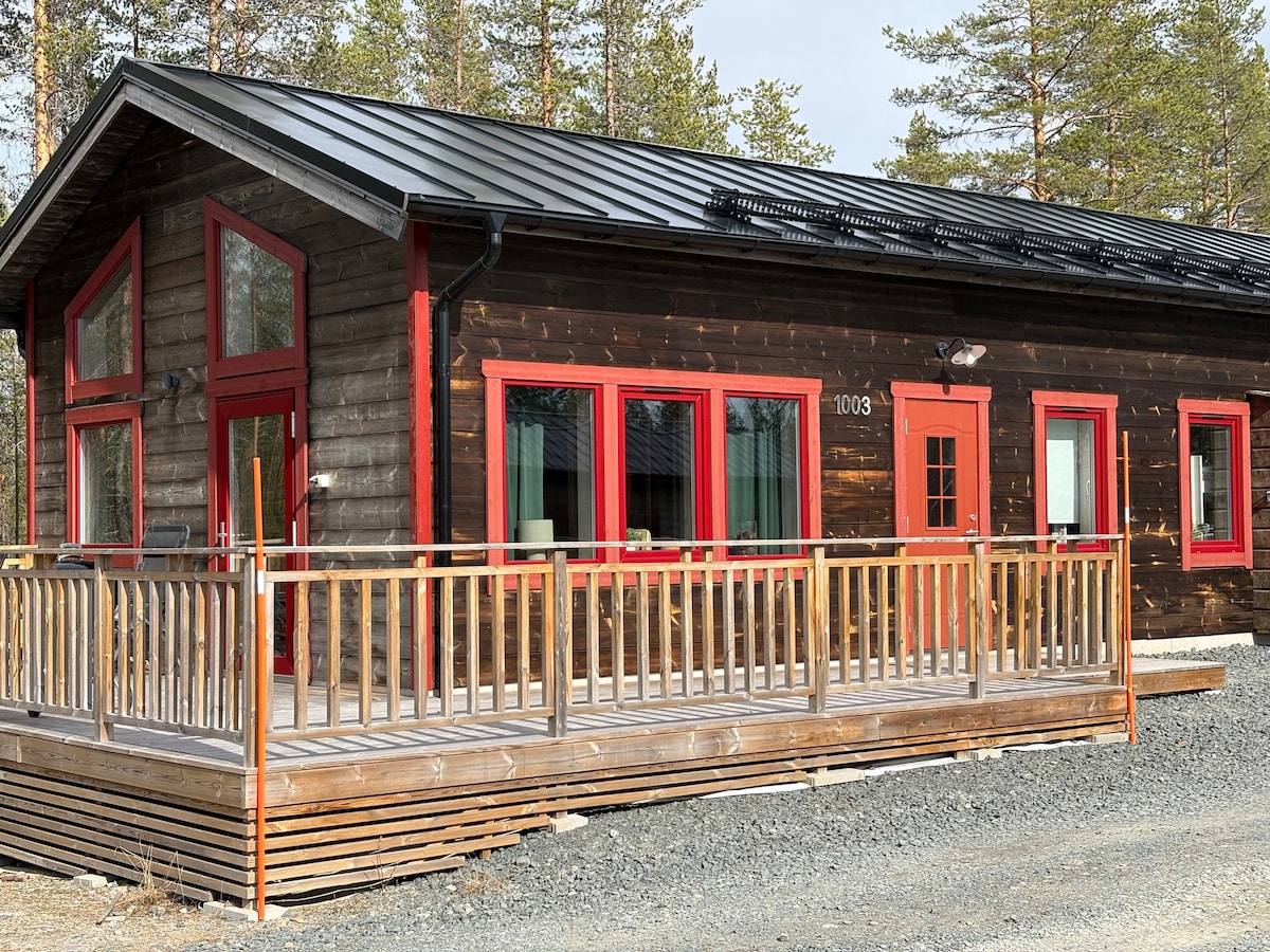 The exterior of the spacious cabin features dark wooden siding, complemented by bright red trim around the windows and doors. A large wooden deck extends from the front, providing an outdoor space with railing. Surrounding conifer trees are visible in the background.