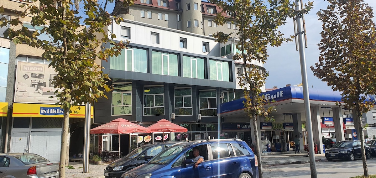 The modern hotel structure features large windows and sleek lines, situated near a gas station and various shops. Outdoor seating areas are visible under awnings, with trees lining the sidewalk. Vehicles are parked along the street, providing a sense of urban activity.