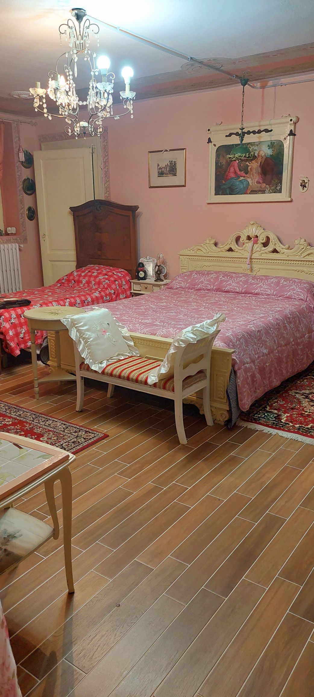 A sleeping area features a spacious bed covered with a pink blanket, complemented by a carved headboard. Two decorative chairs are positioned near the bed, and warm light is provided by a chandelier. The room is adorned with soft pink walls and a patterned rug.