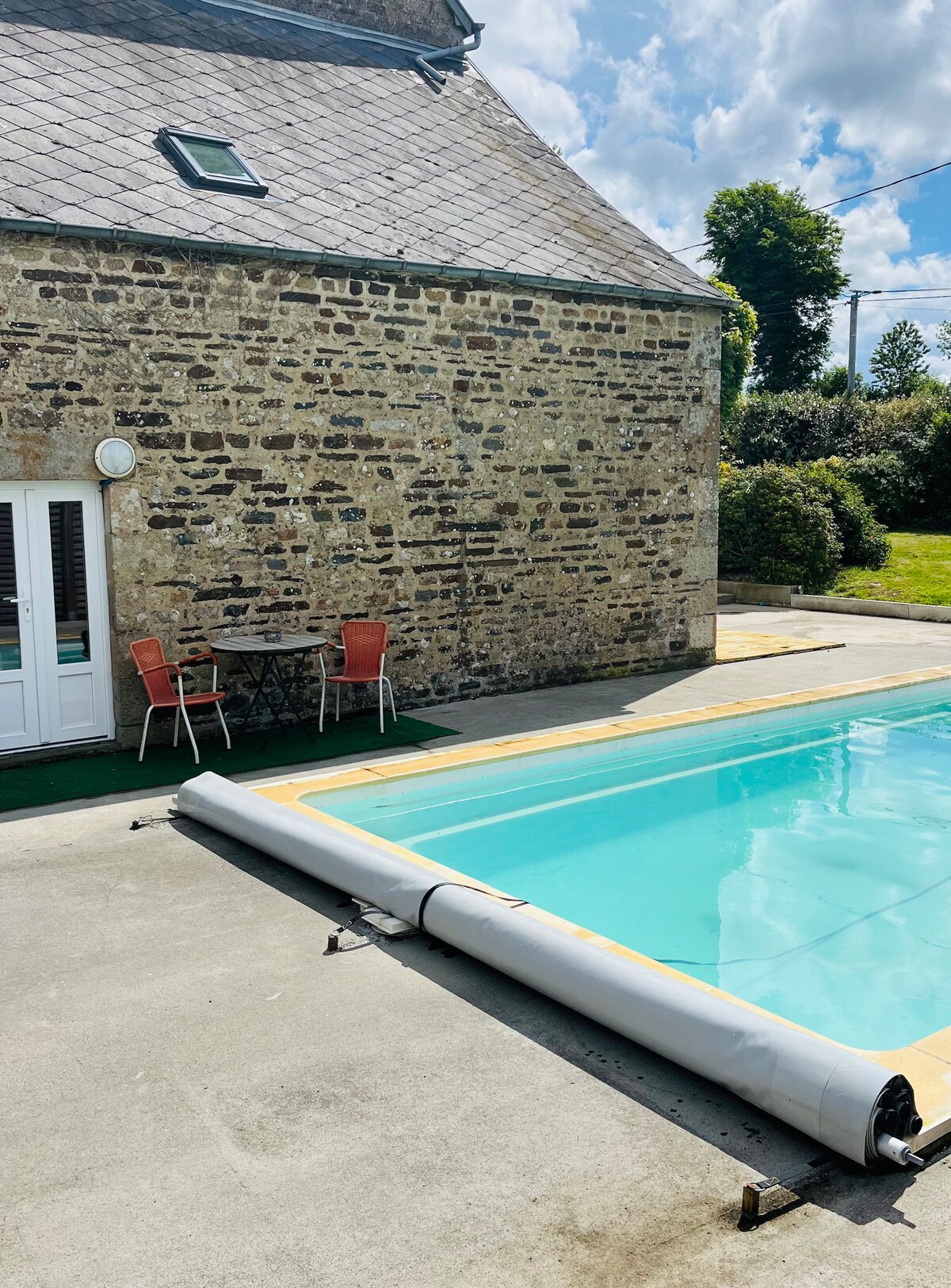 A stone exterior of a rustic house is featured alongside a tranquil swimming pool. Two red chairs and a small black table are situated on the pool deck, providing a space for relaxation. Lush greenery surrounds the area, complementing the outdoor setting.