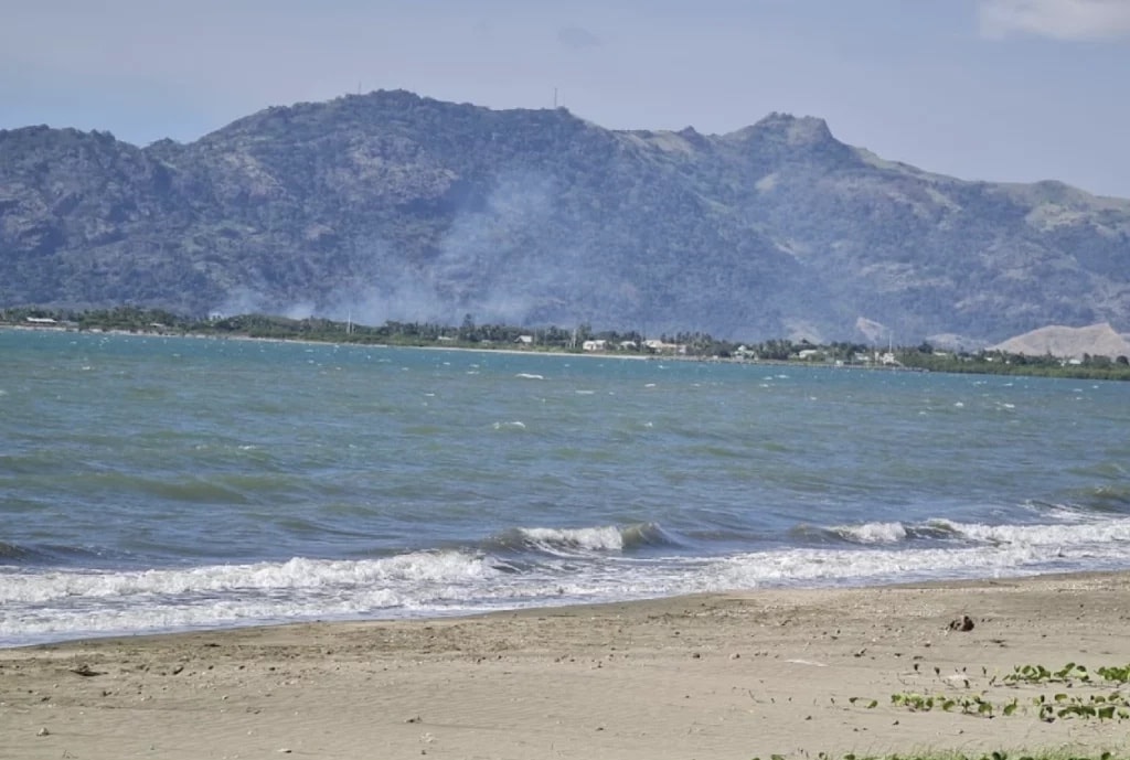 A serene beach scene is presented, with gentle waves lapping against a sandy shore. In the background, a mountain range features rolling hills under a clear sky, creating a peaceful coastal ambiance.