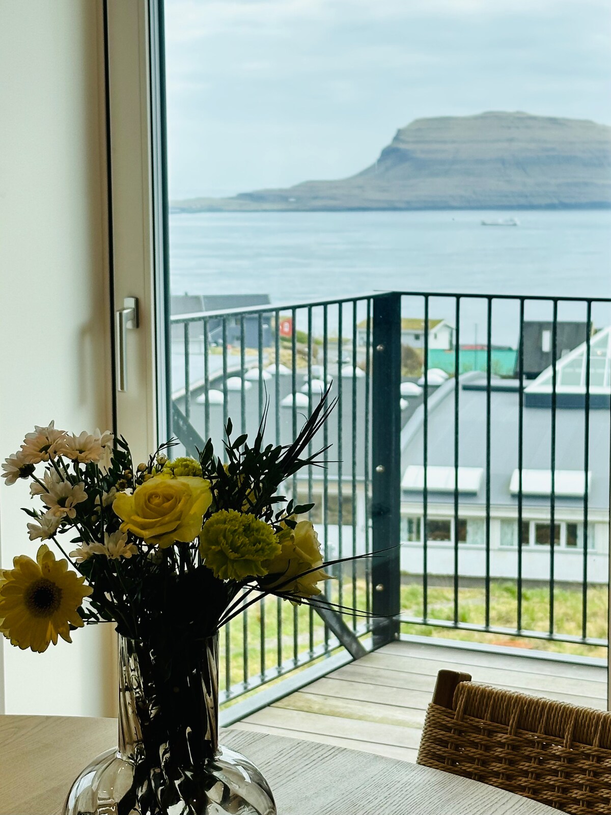A vase filled with fresh flowers including yellow roses and daisies is positioned on a wooden table. In the background, large glass doors frame scenic views of the coastline and a distant mountain, enhancing the openness of the space.
