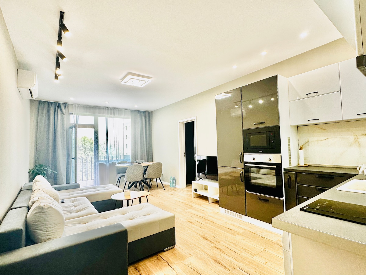 A modern living space features a light-colored sofa and a small circular table, with a dining area visible in the background. Large windows allow natural light to fill the room, complemented by neutral-toned walls and wooden flooring.