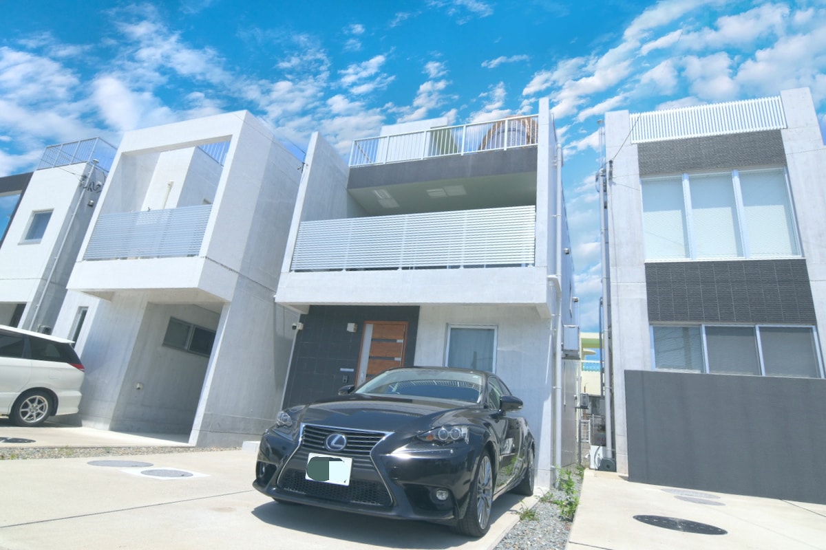 A modern two-story rental property features a sleek exterior design, with a black car parked in front. The building showcases large windows and a balcony on the upper level, surrounded by a clear blue sky dotted with clouds.