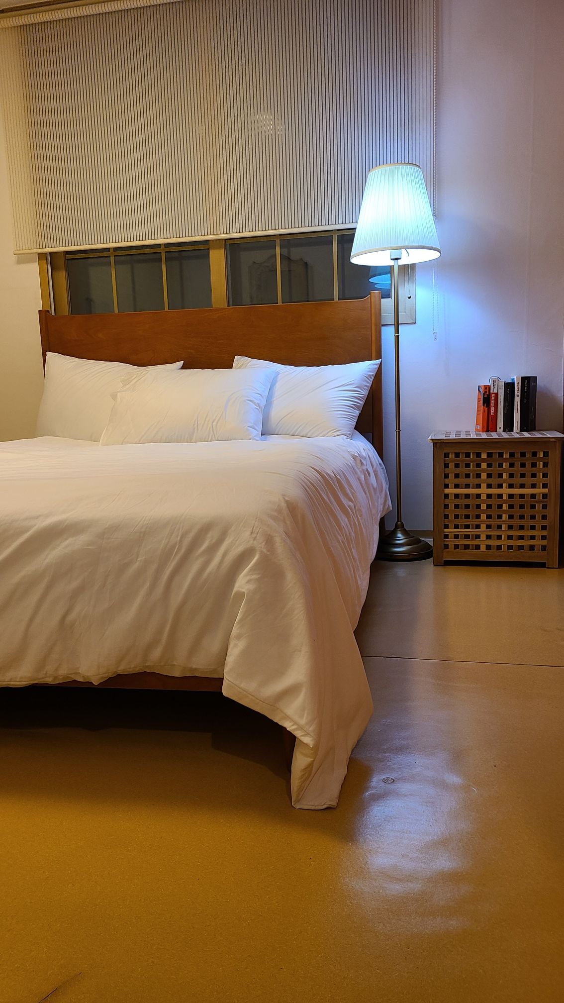 A spacious sleeping area is displayed, featuring a large bed dressed in crisp white linens. A bedside table with a woven design stands beside the bed, accompanied by a reading lamp emitting a soft glow. Books are neatly arranged on the table.