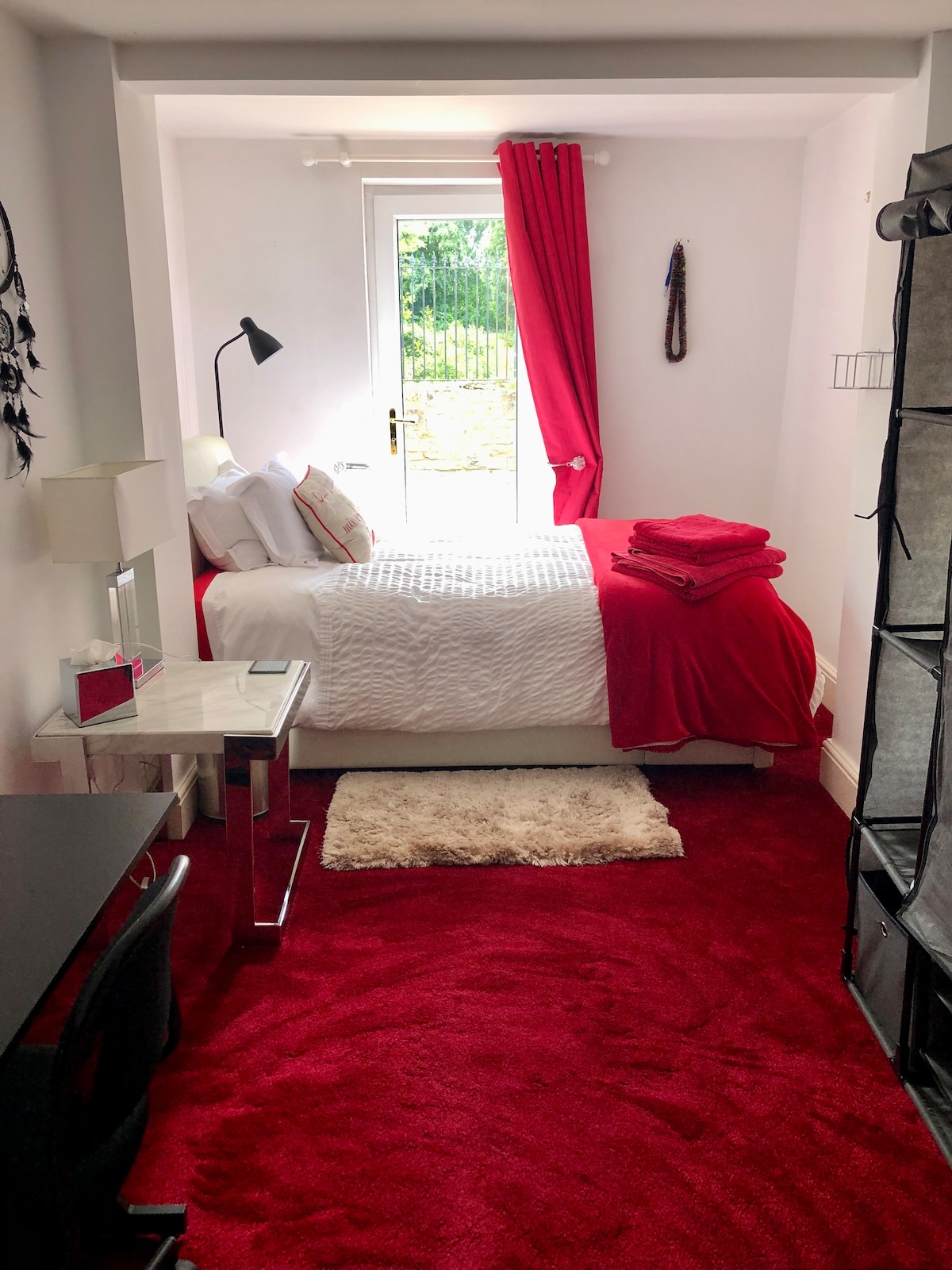 A cozy bedroom features a comfortable bed dressed in white linens and vibrant red accents. Natural light enters through a glass door leading to an outdoor space. A desk and lamp are present, along with a soft rug underfoot, creating a welcoming environment.