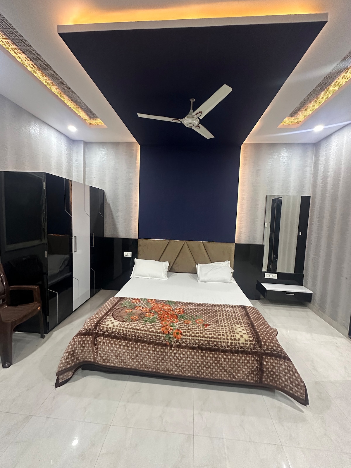 A well-lit sleeping area features a spacious bed dressed with a patterned blanket and decorative accents. A ceiling fan is installed overhead, while mirrored wardrobes and a dressing table are positioned along the walls, providing functional storage and a sense of openness.
