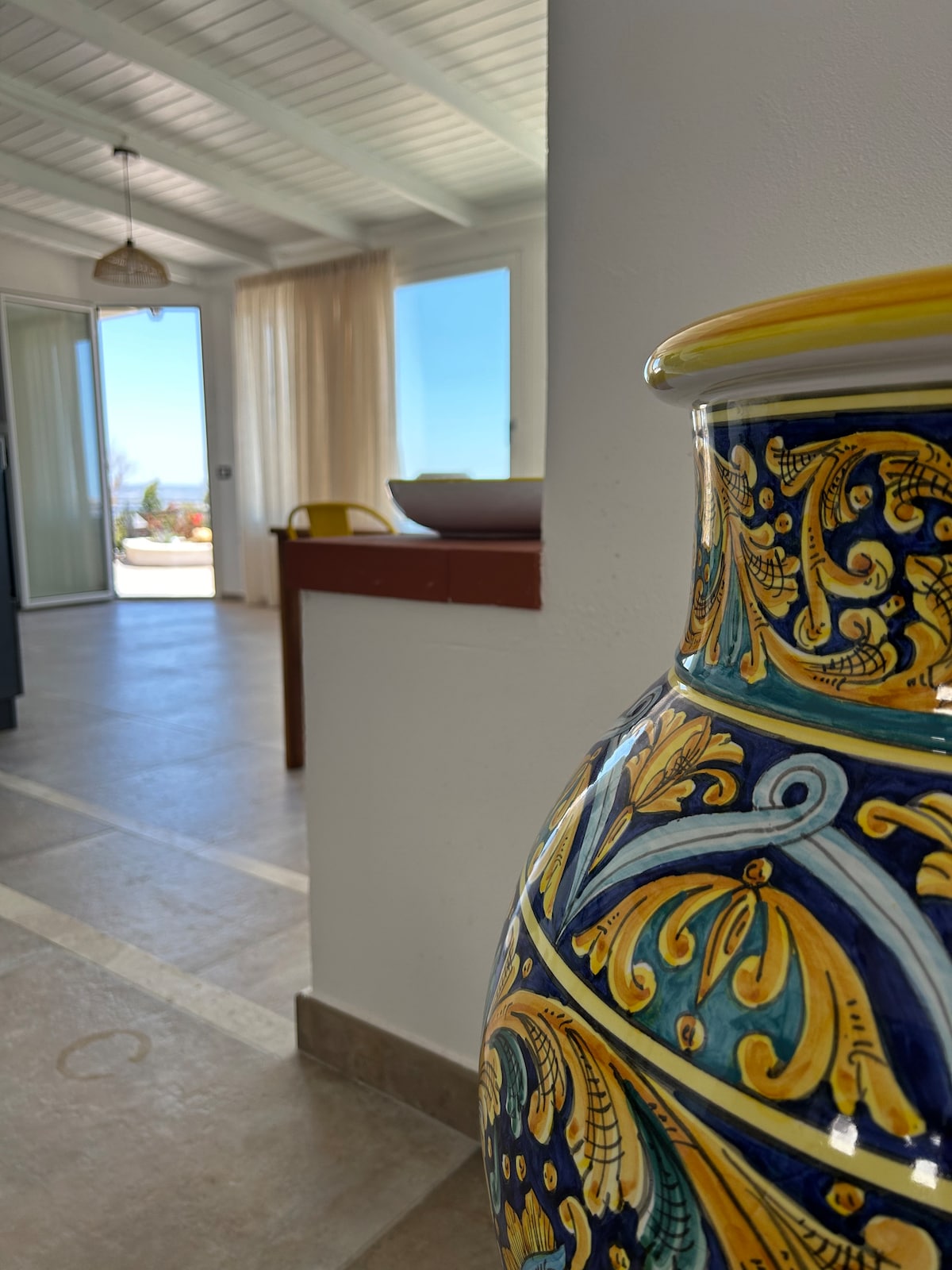 A decorative ceramic vase featuring intricate blue and yellow designs is positioned in the foreground. In the background, a bright and airy space is visible, showcasing large windows that allow natural light and a glimpse of outdoor scenery.