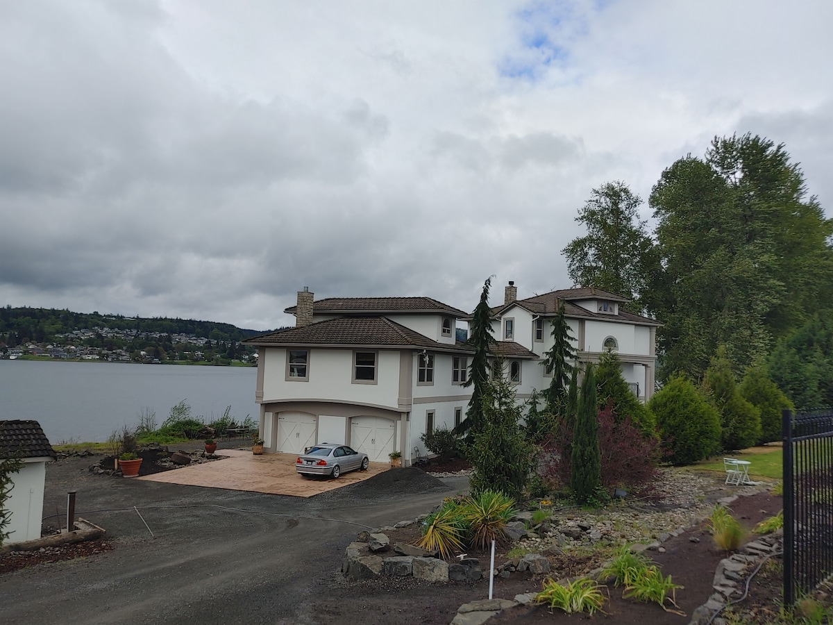 A spacious waterfront property is situated alongside the Columbia River, featuring a two-story design with a mix of stone and stucco exterior. Lush greenery surrounds the house, and a well-maintained driveway leads to the entrance, with trees lining the landscape in the background.