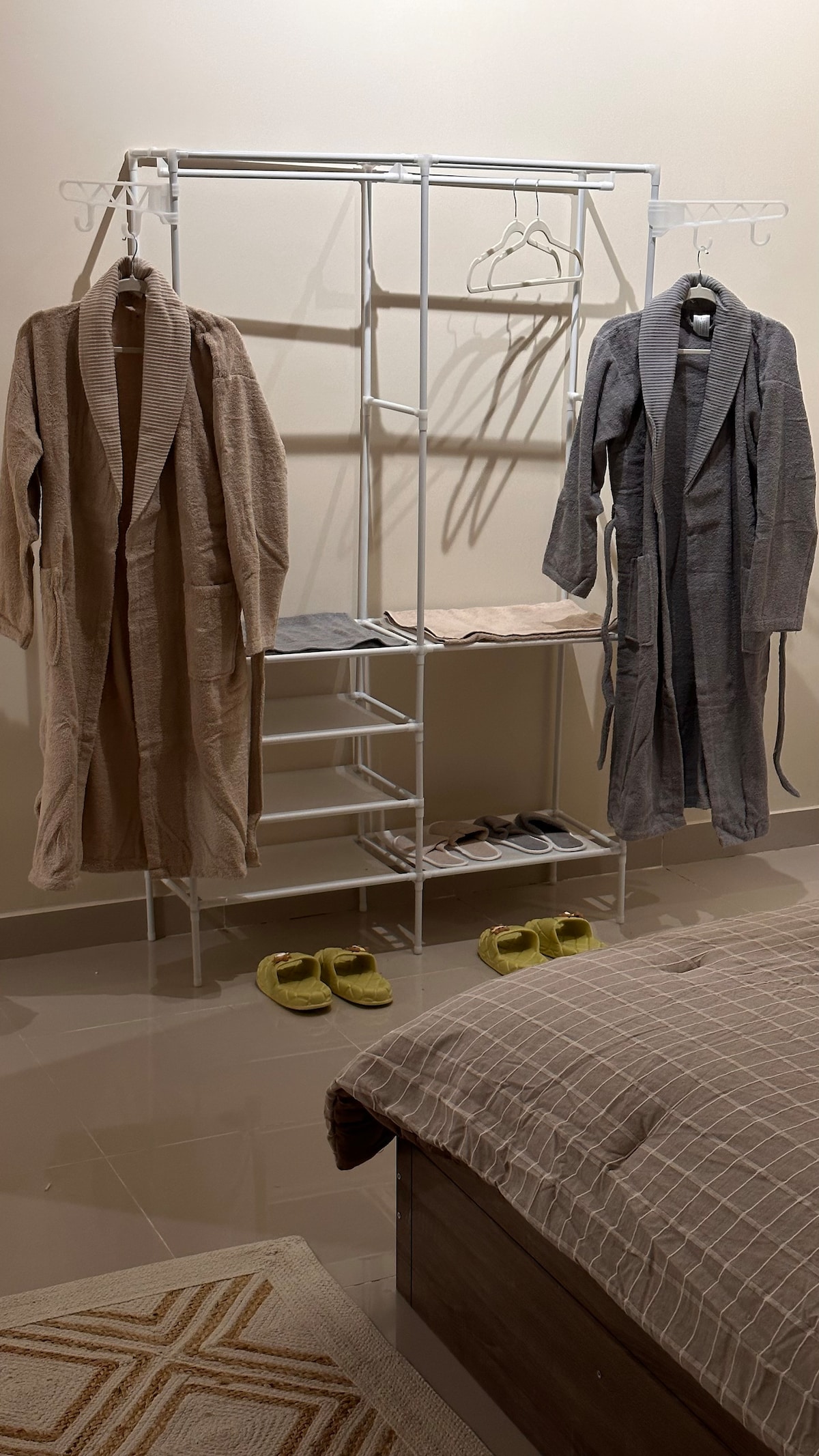 A neatly organized area displays two light-colored robes hanging on a clothing rack. Below, several hangers are available, with a few folded items situated on the shelves. A pair of cozy slippers are placed on the floor next to a bed with a soft, checkered blanket.
