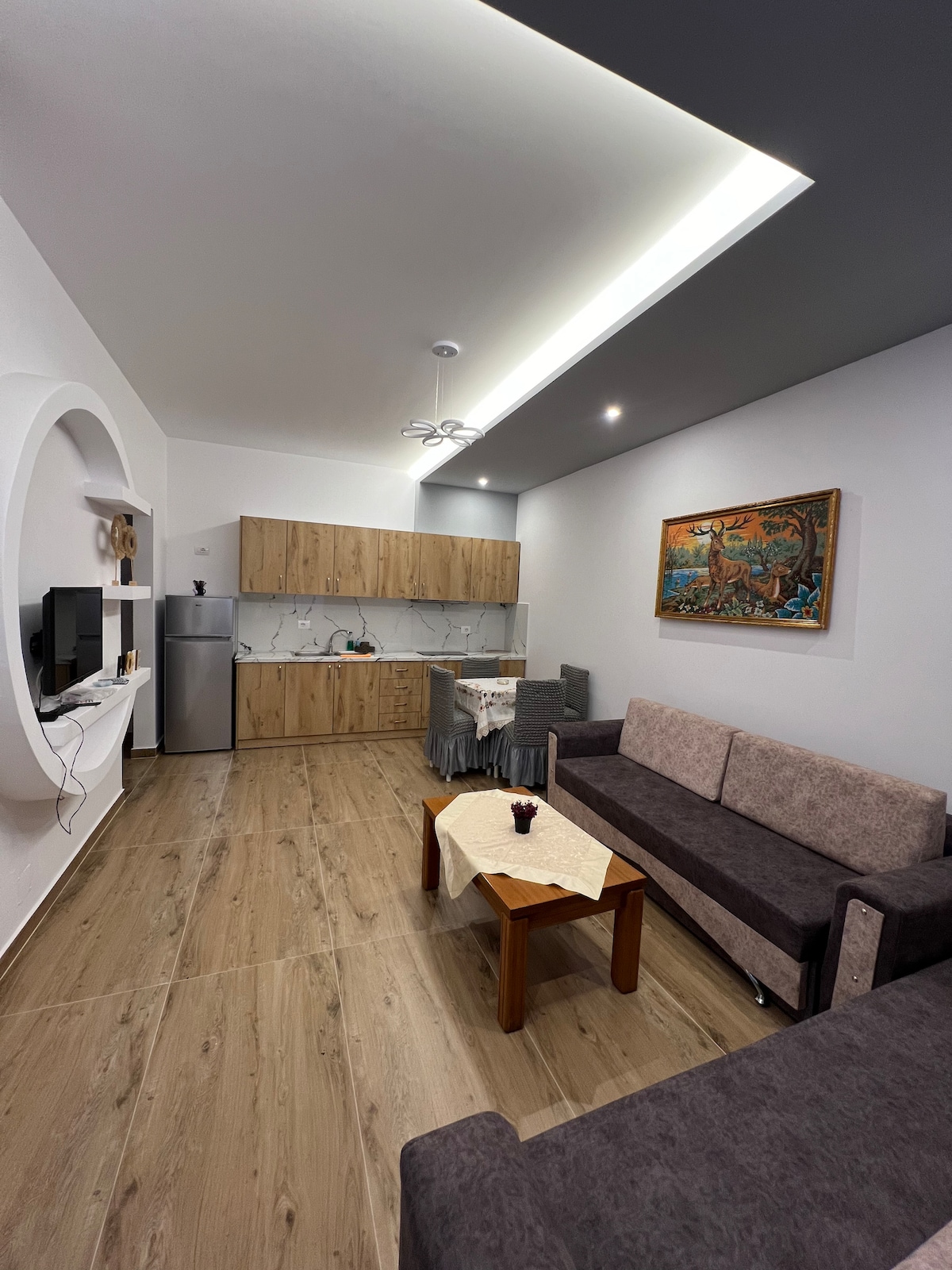 A bright living space features light wood cabinetry integrated with a modern kitchen. A cozy seating area includes two gray sofas surrounding a wooden coffee table. A wall-mounted television complements the room, while artwork adds color above the dining setup in the background.