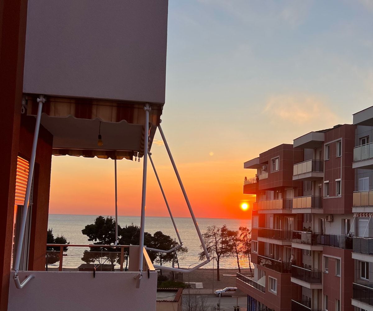 A sunset view is captured from a balcony, with the horizon displaying vibrant orange and pink hues. The calm sea reflects the fading light, while nearby trees and buildings frame the picturesque coastal landscape.