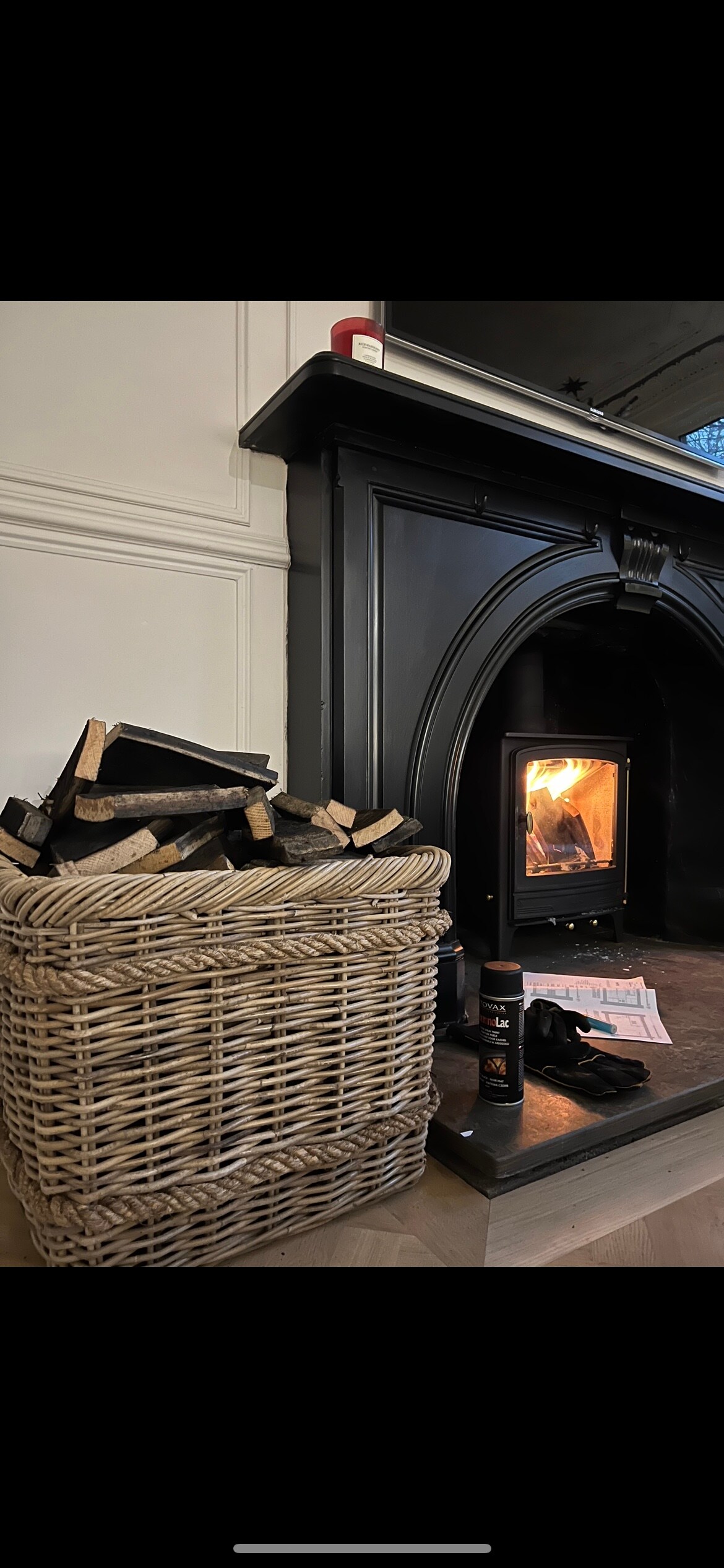 A wicker basket filled with neatly stacked firewood is positioned next to a black fireplace. A warm flame is visible within the fireplace, creating a cozy focal point in the room. Natural light filters in, enhancing the inviting atmosphere.