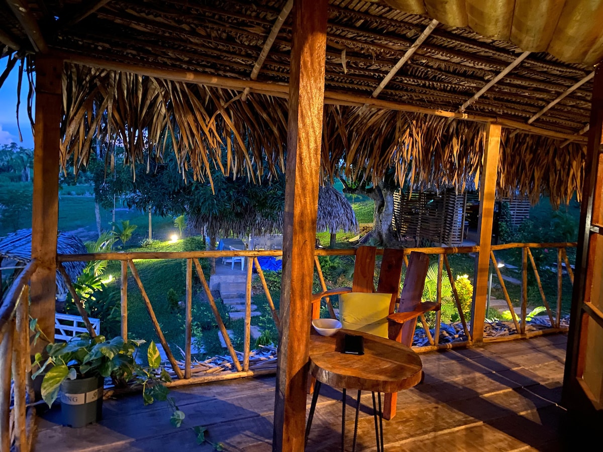 A covered porch offers a view of the lush landscape, framed by wooden beams and a thatched roof. An inviting wooden chair and small table are positioned for relaxation, while soft lighting illuminates the surrounding greenery.