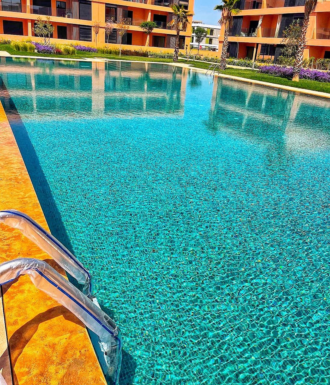 A large, inviting swimming pool features clear, turquoise water surrounded by manicured gardens. Fresh greenery and blooming flowers are visible in the background, complemented by modern buildings. Steps lead into the pool, highlighting the serene atmosphere of the residence.