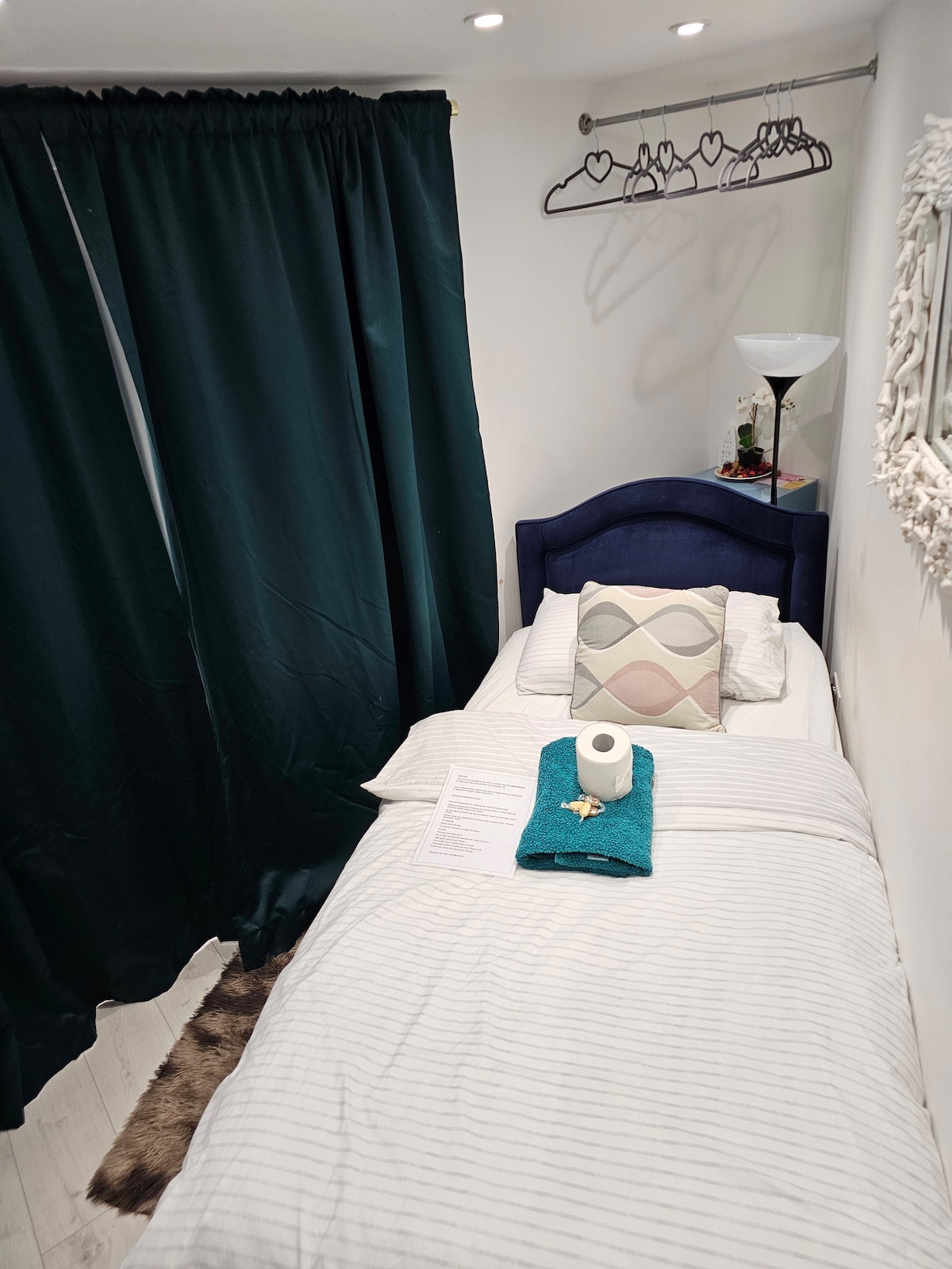 A cozy bedroom features a narrow bed dressed in white linens with a decorative throw and a rolled towel placed on top. A tall lamp illuminates the space, while dark green curtains provide privacy. A hanger bar is visible above the bed, adding functionality.