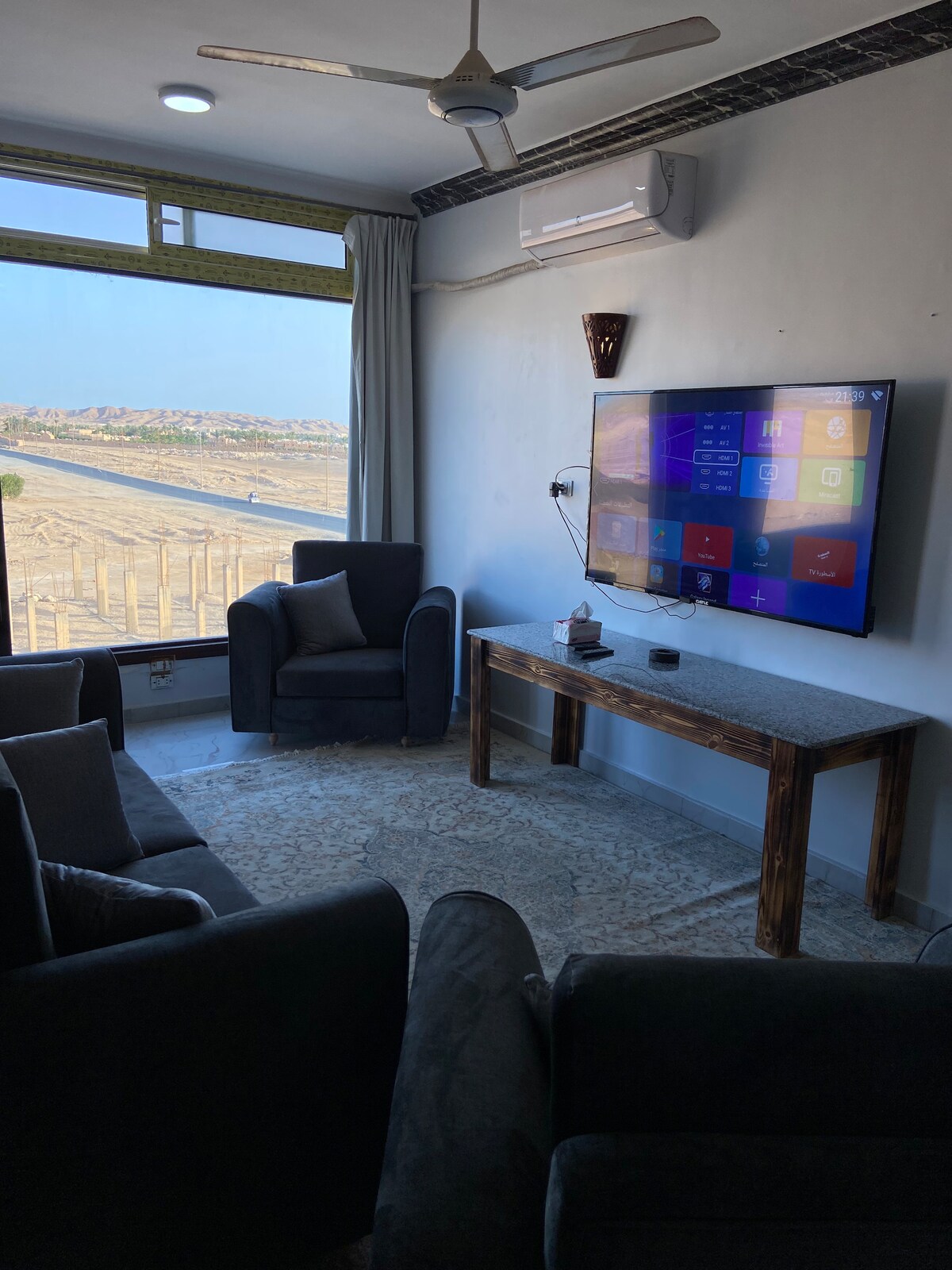 A comfortable living area is depicted, featuring two armchairs and a contemporary sofa arranged around a wooden coffee table. A flat-screen TV is mounted on the wall, with a view of the scenery visible through a large window. Light curtains frame the window.