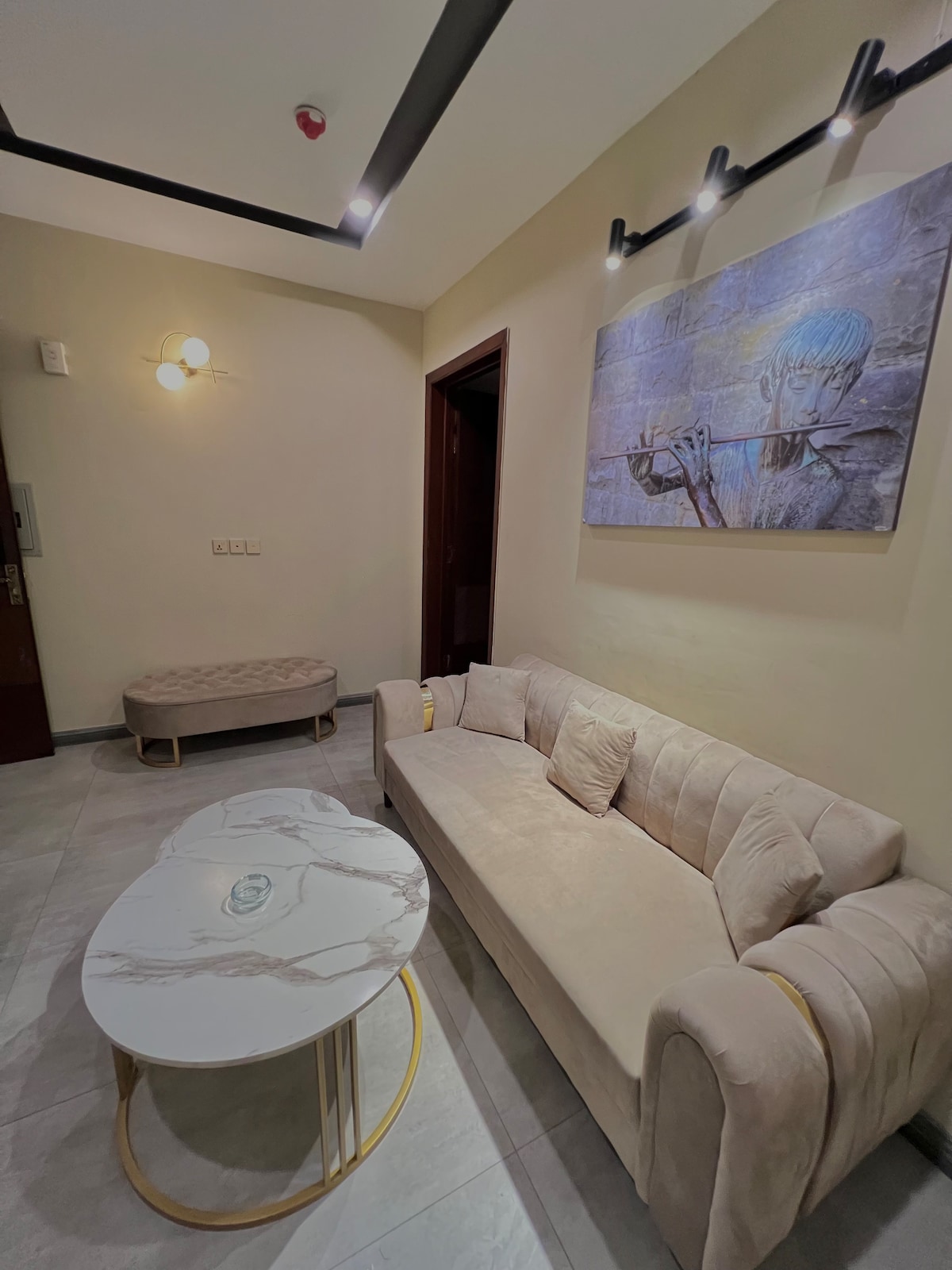 A light-colored sofa with curved edges is positioned near a marble coffee table featuring gold accents. Above, a wall art piece depicts a figure playing the flute, adding character to the room. Soft lighting enhances the serene atmosphere.