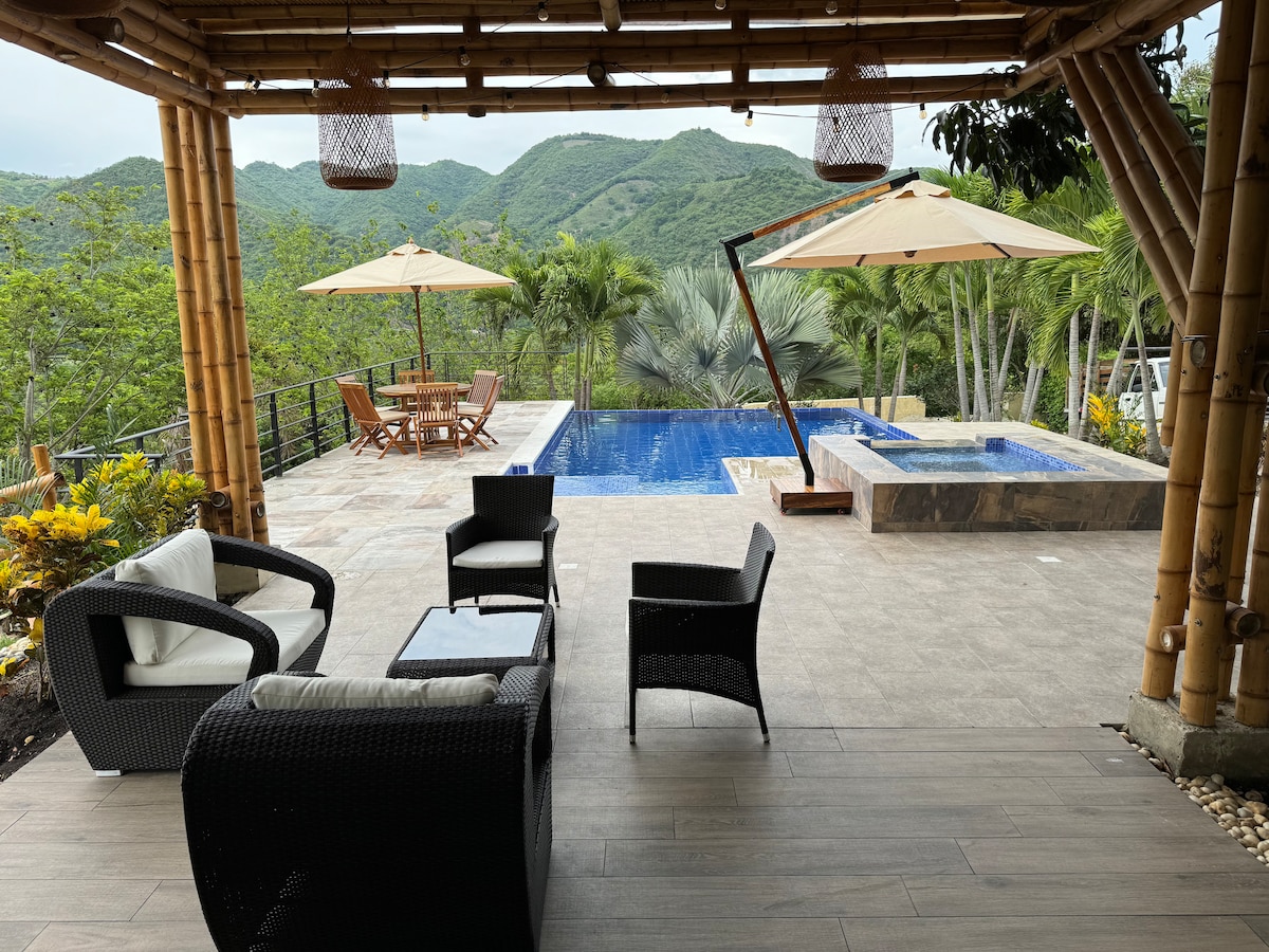 A spacious patio area features comfortable seating arrangements, including two armchairs and a small table. In the background, a swimming pool and jacuzzi are visible, surrounded by lush greenery and mountains. Two umbrellas provide shade over a dining table situated nearby.