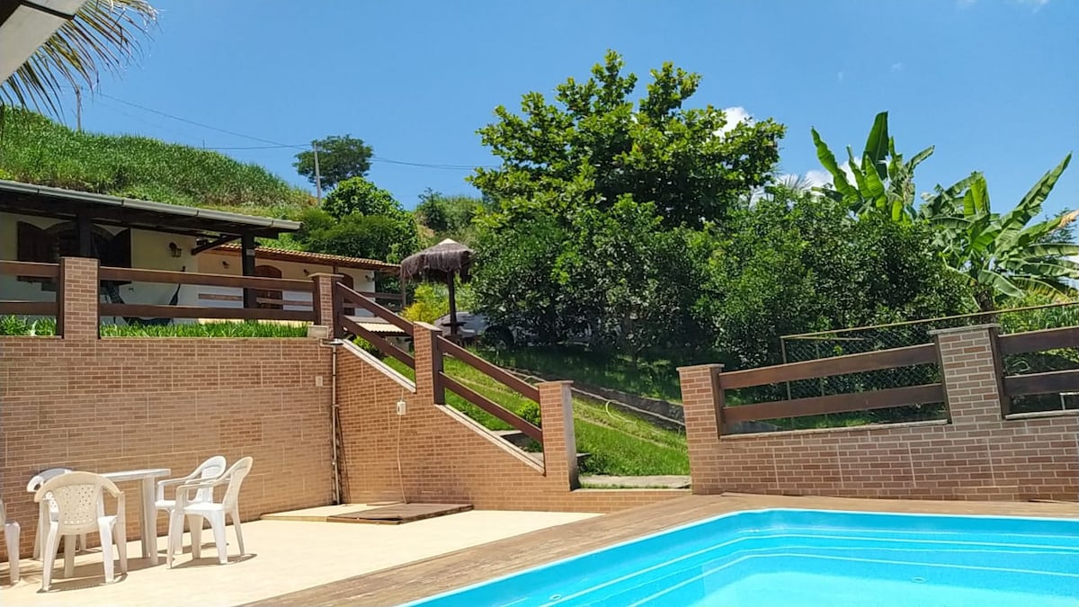 A tranquil outdoor area features a clear pool surrounded by a patio. White chairs and a table are situated to one side, with steps leading to a grassy hill adorned with trees and plants, creating a serene environment.