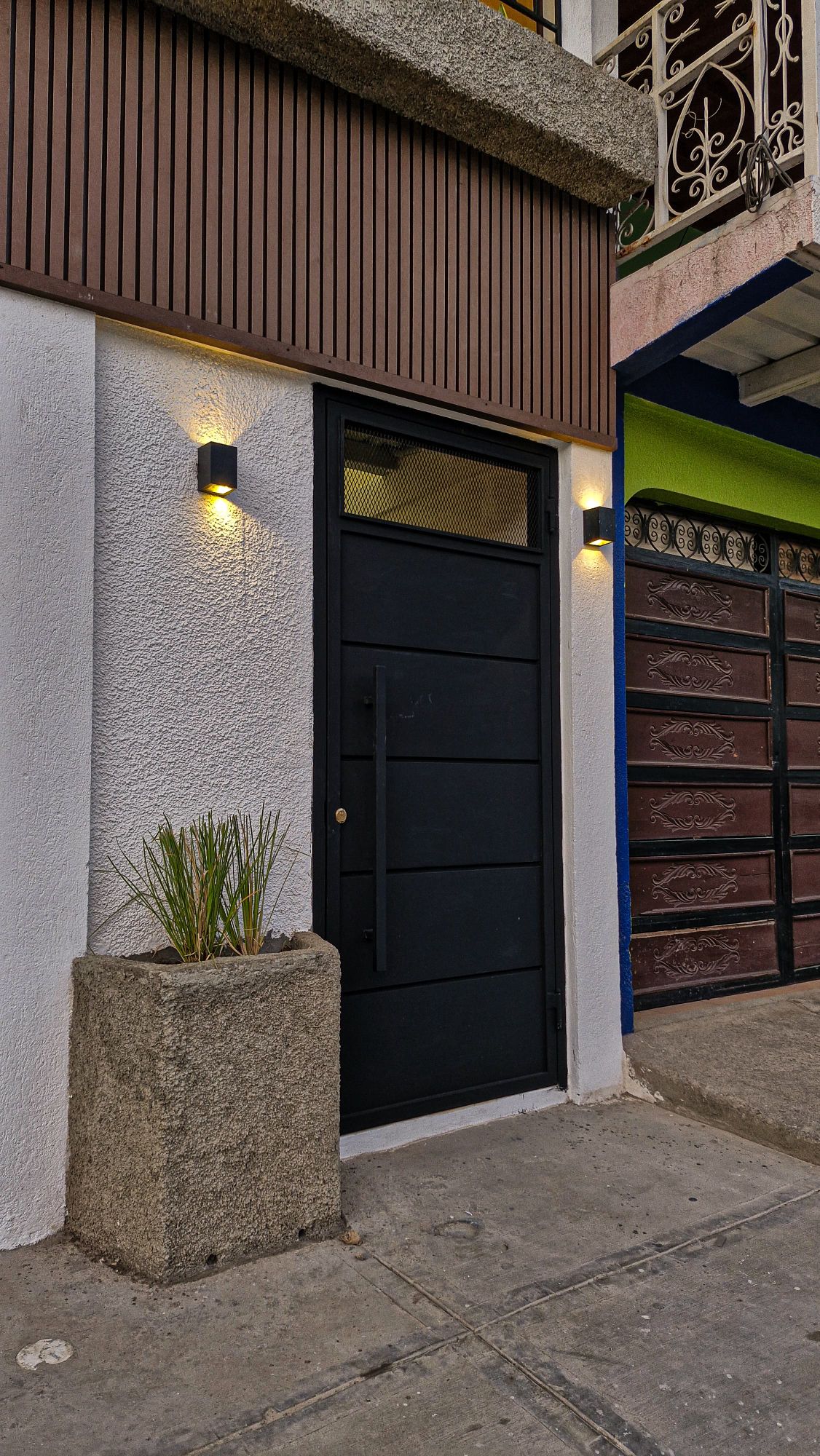 The entrance to the accommodation features a modern black door with vertical panels, flanked by two wall-mounted lights providing soft illumination. A simple planter with greenery adds a touch of nature to the concrete base, situated on a well-maintained sidewalk.