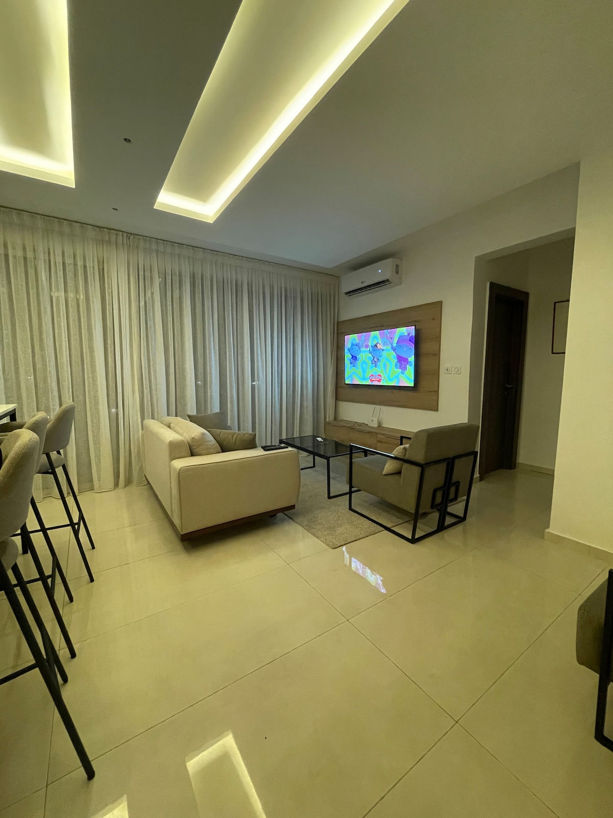 A modern living area features a spacious sofa and two armchairs, arranged around a coffee table. A flat-screen television is mounted on the wall, and large windows are dressed with sheer curtains, allowing natural light to fill the room.